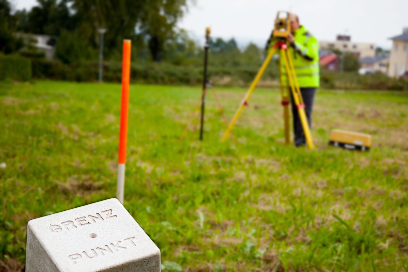 Grenzpunkt-Stein auf einer Wiese, dahinter ein Vermessungstechniker am Tachymeter